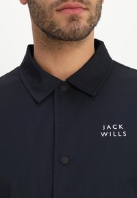 Detailní záběr dolní části obličeje a krku muže, který má na sobě černou bundu s špičatým límcem a bílým logem "Jack Wills" na hrudi.