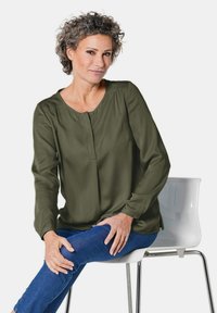Olivegrüne Bluse mit langen Ärmeln und Rundhalsausschnitt, mit einer Knopfdetail vorne. Kombiniert mit blauen Jeans.