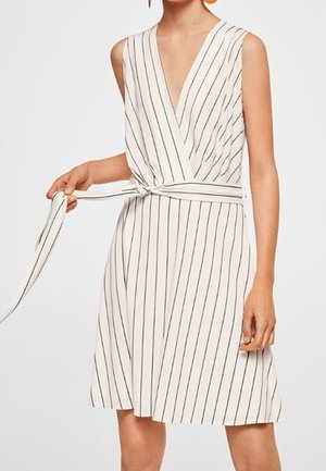 Robe blanche sans manches avec des rayures verticales noires, un décolleté en V profond, et une ceinture nouée à la taille tenue par une personne ajustant le nœud.