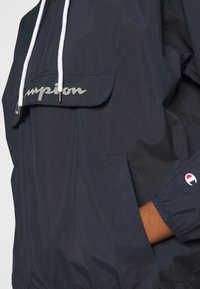 Marineblaue Windjacke mit einer Fronttasche, weißen Kordeln und einem reflektierenden "Champion"-Logo. Verfügt über elastische Bündchen und eine weiche Textur.