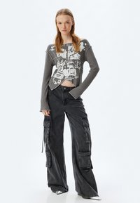 Grauer gerippter Cropped-Top mit Grafikdruck; kombiniert mit locker sitzenden schwarzen Cargo-Hosen, die mehrere Taschen und Kordelzugdetails aufweisen.