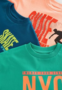 Trois t-shirts col rond : teal, marine et pêche. Chacun arbore des motifs imprimés distinctifs dans des couleurs contrastées. Texture en coton doux.