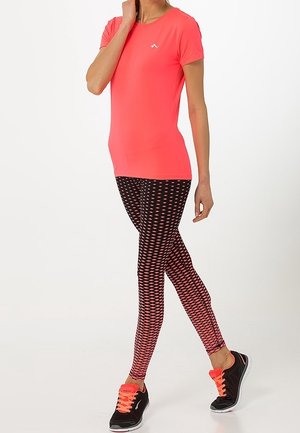 Mujer con camisa de manga corta color coral brillante, leggings negros con patrón de diamantes rosas y zapatillas negras con cordones coral, de pie con una pierna cruzada.