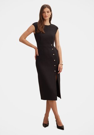 Femme debout portant une robe midi noire cintrée avec des manches courtes, des boutons en forme de cœur sur le côté, une fente à la cuisse, des talons noirs et des boucles d'oreilles créoles dorées.