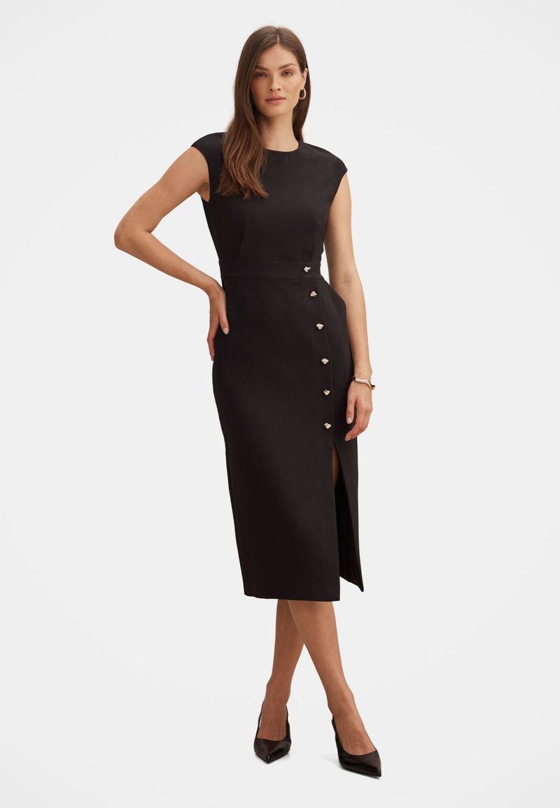 Femme debout portant une robe midi noire cintrée avec des manches courtes, des boutons en forme de cœur sur le côté, une fente à la cuisse, des talons noirs et des boucles d'oreilles créoles dorées.