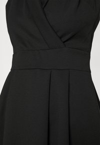 Detailní záběr černých šatů s širokým pasem, skládáním sukně a překrývajícím se živůtkem ve tvaru V.