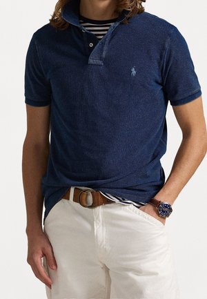 Polo bleu marine avec col, manches courtes et un logo sur la poitrine. Associé à un short blanc et une ceinture marron.