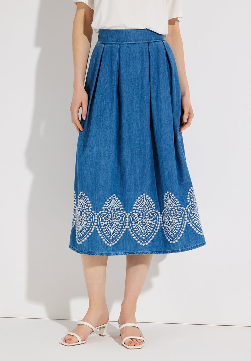 Frau trägt einen blauen Midirock mit weißer geometrischer Stickerei am Saum und weiße Sandalen mit Absatz, steht vor einem weißen Hintergrund.