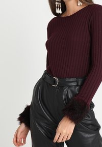Vrouw in een geribbelde bordeauxrode langemouwen top met zwarte nepbont manchetten, gecombineerd met hoge zwarte leren broeken en een riem.