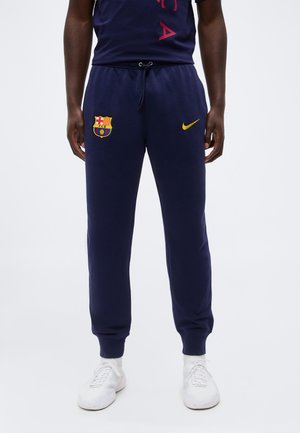 Homme portant un pantalon de sport bleu marine avec l’écusson du FC Barcelone sur la cuisse gauche et le logo jaune Nike sur la cuisse droite, assorti à des baskets blanches.