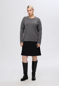 Pelēka adīta džemperis ar apaļu kakla izgriezumu, apvienots ar melnu ceļgala garuma svārku. Melni ceļgala garuma zābaki papildina apģērbu.