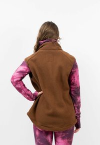 Bruine fleece vest met hoge kraag, mouwloos ontwerp en een gestructureerde afwerking. Draagt over een paarse tie-dye longsleeve en leggings.