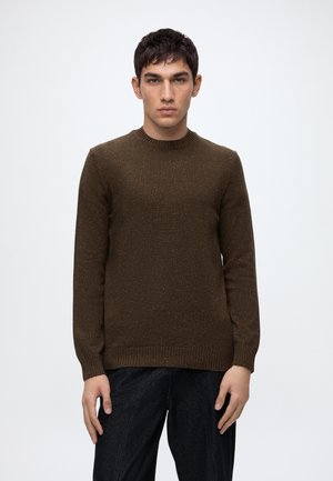 Jeune homme aux cheveux courts et foncés, portant un pull en tricot marron et un pantalon foncé, debout devant un fond clair uni.