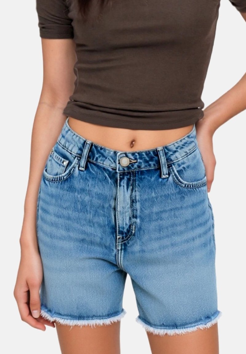 Femme portant un short en denim bleu délavé taille haute avec des ourlets effilochés et un t-shirt marron court, debout avec une main sur la hanche.