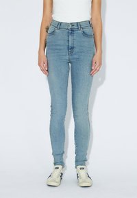 Persona vestida con jeans skinny de color azul claro, top blanco sin mangas y zapatillas blancas con rayas azules, de pie contra un fondo blanco liso.