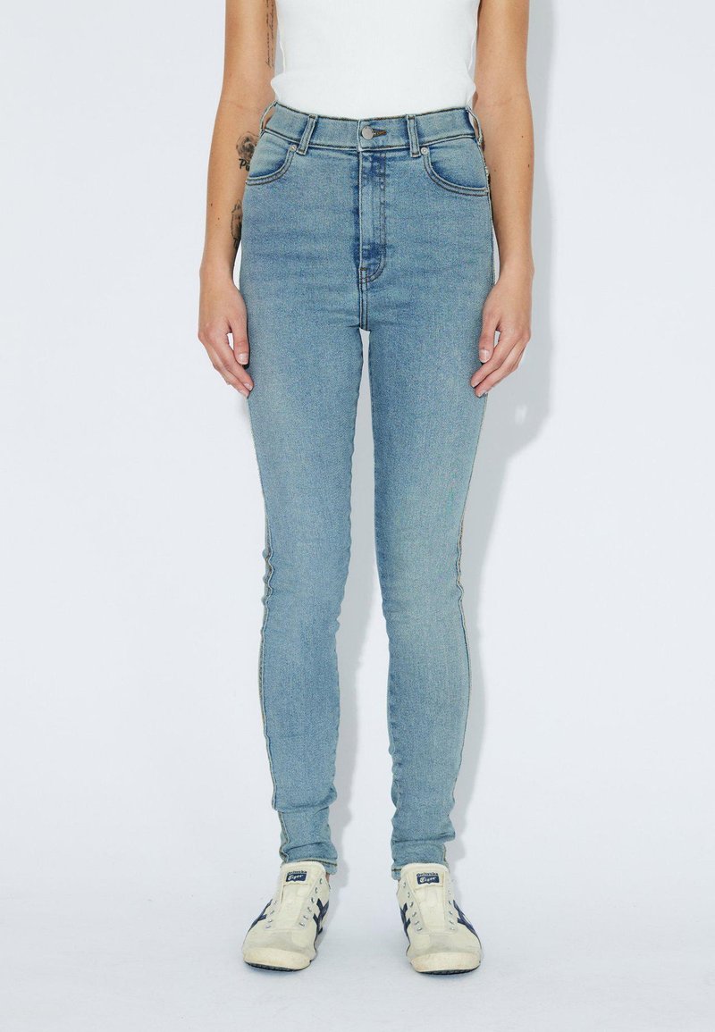 Persoon in lichtblauwe skinny jeans, een witte tanktop en witte sneakers met blauwe strepen, staand voor een effen witte achtergrond.