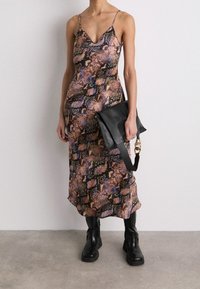 Femme portant une robe nuisette à imprimé serpent multicolore avec des bretelles fines, des bottes noires à semelle épaisse et tenant une pochette en cuir noir.