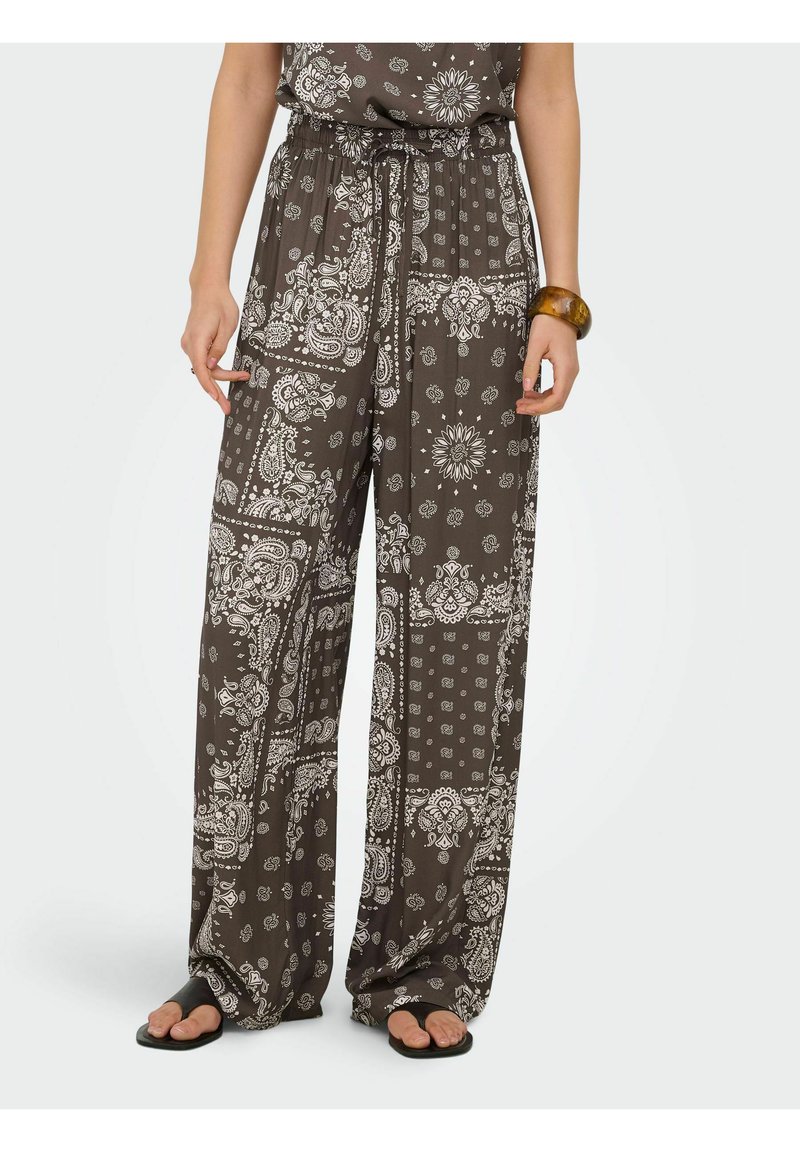 Pantalon marron à jambes larges avec motifs paisley et floraux blancs, taille élastique avec cordon, porté avec des sandales noires à bouts ouverts.