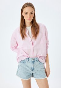 Camisa de botones de color rosa claro con mangas largas, combinada con pantalones cortos de denim azul con tachuelas plateadas y dobladillo deshilachado. Detalle de bolsillo sencillo.