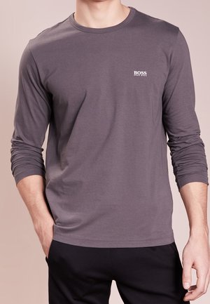 Mann trägt ein graues Langarmshirt von Hugo Boss, die Hände in den Taschen, vor einem schlichten beigen Hintergrund.