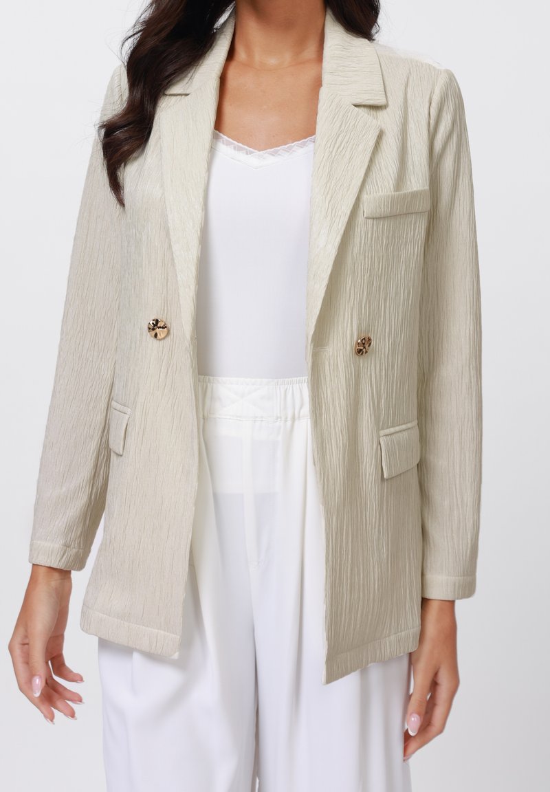 Femme portant un blazer beige clair texturé avec des boutons dorés sur un haut blanc et un pantalon blanc taille haute, sur un fond uni.