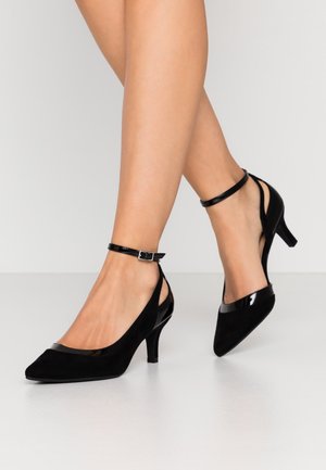 Jambes portant des escarpins en daim noir à bout pointu avec brides à la cheville, talon aiguille de hauteur moyenne, posées sur un sol lisse gris clair.