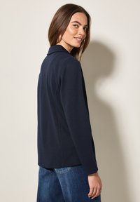 Veste bleu marine avec un col, design ajusté, texture douce et manches longues, associée à un jean en denim.