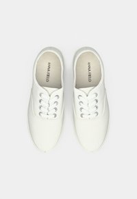 Hvide kanvas sneakers med sølvøjer, flade snørebånd, rund tå og en syet søm langs sålen. Indeni er mærkenavnet "ANNA FIELD."