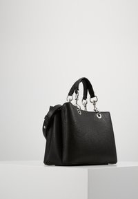 Musta tekstuuripöksi käekott kahe lühikese sangaga, hõbedaste detailidega ja eemaldatava õlarihmaga valgel pedestalil.