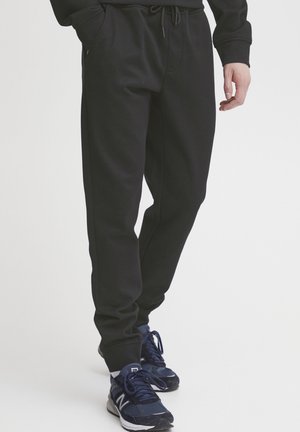 Bas du corps d'une personne portant un pantalon de jogging noir et des baskets bleu marine avec des semelles blanches, debout devant un fond clair uni.