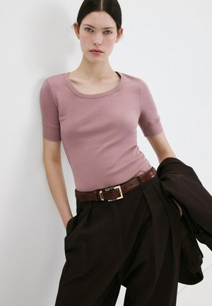 Femme aux cheveux foncés portant un haut ajusté à manches courtes rose, un pantalon noir taille haute avec une ceinture marron, tenant une veste noire, debout contre un mur blanc.