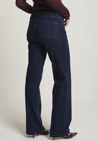 Person klädd i mörkblå vida jeans, vinröd topp och vinröda spetsiga klackar, stående mot en enkel ljus bakgrund.