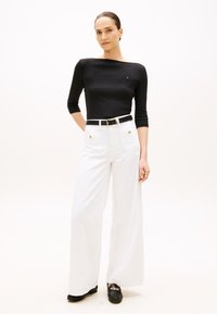 Crna rebrasta majica dugih rukava, bijele široke hlače visokog struka s zlatnim gumbima, crni remen i crne mokasine s lančanim detaljem.