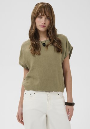 Žena s dugom smeđom kosom koja nosi maslinasto zelenu bluzu kratkih rukava, bijele hlače, crni ogrlicu i crnu narukvicu, na jednobojnoj pozadini.