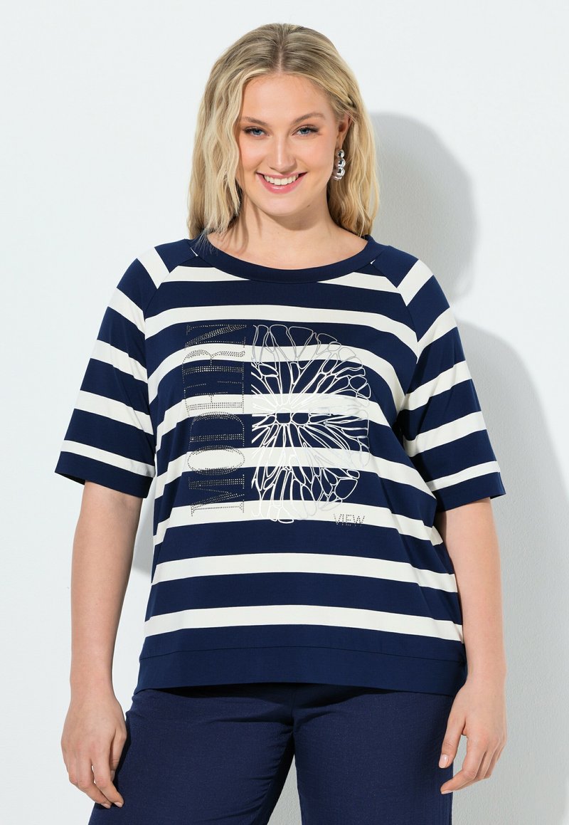 Usměvavá žena s vlnitými blond vlasy nosí tmavě modré a bílé pruhované tričko s krátkými rukávy s květinovým a textovým motivem, doplněné tmavě modrými kalhotami.