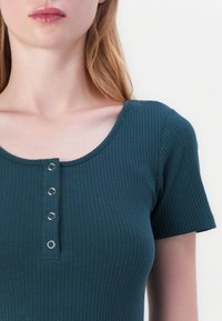 Žebrovaný, tyrkysový top s krátkým rukávem a kulatým výstřihem, se zapínáním na pět knoflíků s kovovými očky. Měkká textura, přiléhavý střih.
