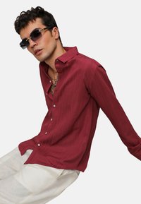 Camicia a maniche lunghe bordeaux con sottili righe verticali, dotata di bottoni sul davanti e colletto, abbinata a pantaloni larghi di colore chiaro.