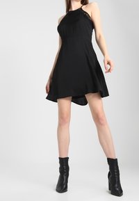 Black halter neck dress with lace accents, a flowing skirt, and asymmetrical hem. Model wears black ankle boots. Smooth texture.