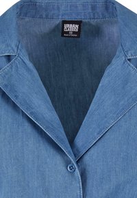 Col de chemise en denim avec un bouton bleu unique, coutures visibles, et une étiquette de vêtement noire indiquant "Urban Classics XS Fabriqué au Pakistan".