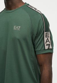 Κοντινό πλάνο ενός ατόμου που φοράει πράσινο T-shirt EA7 Emporio Armani με λογότυπο στο στήθος και ριγέ με επωνυμία στα μανίκια.