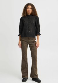 Camicia nera con bottoni, motivo testurizzato, polsini arricciati e colletto alto, abbinata a pantaloni marroni a zampa d'elefante e scarpe nere.