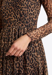 Leopardi-kuosiset mekko, jossa on läpinäkyvät pitkät hihat, joustavat hihansuut, joissa on aaltoilevat reuna, ja laskostettu hame ruskealla ja mustalla sävyllä.