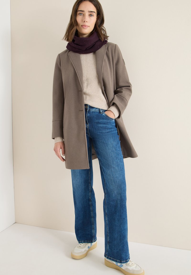 Manteau marron, pull beige clair, jean bleu à jambes larges et baskets à motifs. Écharpe violette foncée avec des points subtils. Textures lisses.