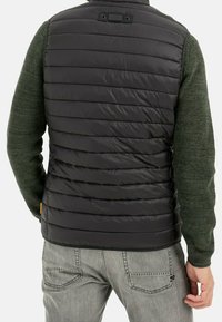 Hombre con chaleco acolchado negro sobre suéter de punto verde y jeans gris claro, visto desde atrás.