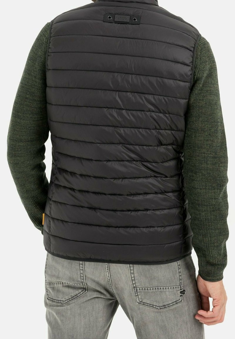 Hombre con chaleco acolchado negro sobre suéter de punto verde y jeans gris claro, visto desde atrás.