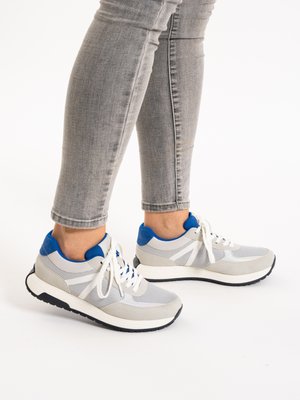 Person wearing gray jeans and gray sneakers with blue accents, white laces, and black soles, standing on a white surface.