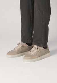 Zapatillas beige elaboradas con materiales de cuero suave y gamuza, con puntera redonda, suelas blancas planas y cordones, combinadas con pantalones gris oscuro.