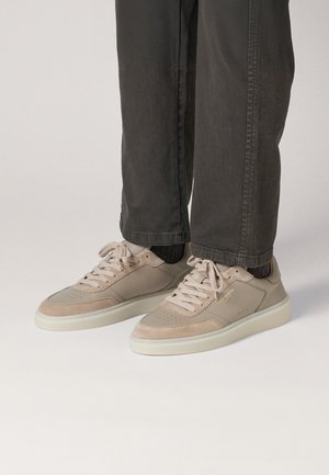 Beige sneakers met glad leer en suede materialen, ronde neus, platte witte zolen en veters, gecombineerd met donkergrijze broek.