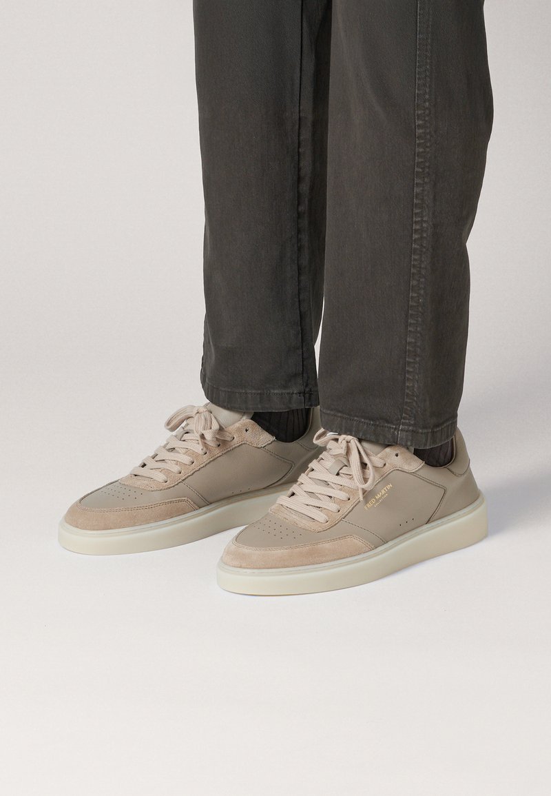 Zapatillas beige elaboradas con materiales de cuero suave y gamuza, con puntera redonda, suelas blancas planas y cordones, combinadas con pantalones gris oscuro.