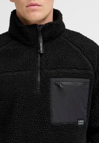 Black fleece pullover with high collar, half-zip front, and a contrasting black zippered pocket on the chest. Soft, textured fabric.